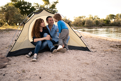 happy family in tent at campsite, father, mother and little son laughteen to something. camping, tourism, hike and people concept