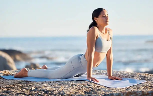 Photo of Yoga, fitness and woman at the beach for exercise while in cobra pose for breathing, zen and wellness on rock while on vacation in summer. Female by sea to meditate, workout and practice mindfulness