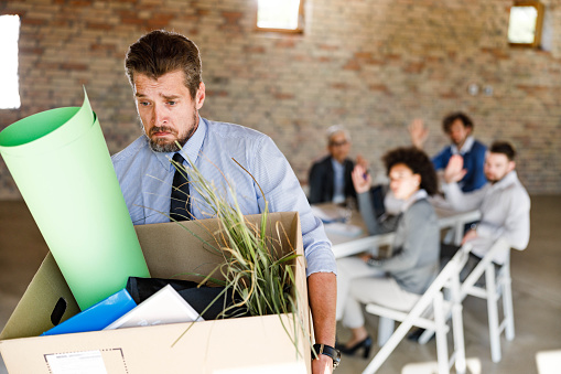 Sad entrepreneur carrying his belongings while leaving the office after being fired. His colleagues are in the background.