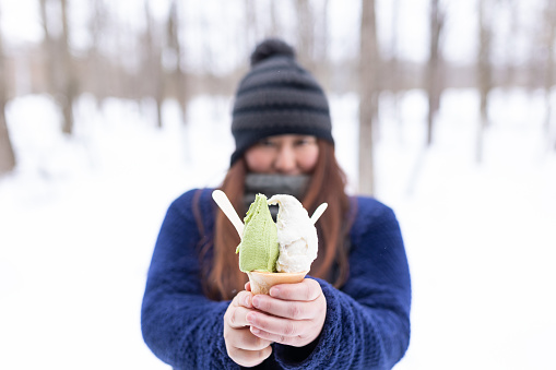 A winter forest scene with a Japanese woman eating ice cream with matcha and rum raisin flavors in a cone.