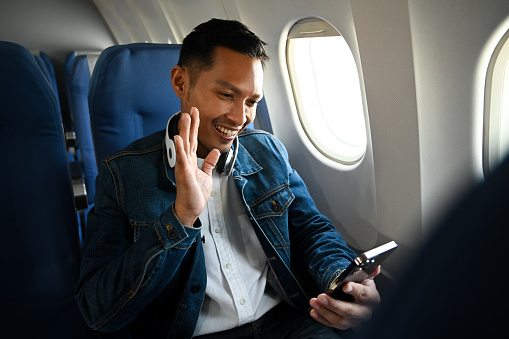 Smiling young hipster male passengers using smart phone having video call on mobile phone while sitting on aircraft cabin.