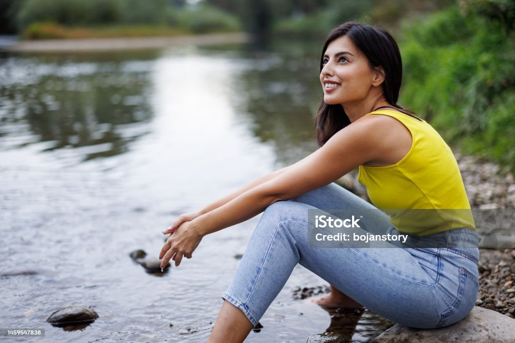 Pure enjoyment Young woman sitting on rock by a stream with bare feet in water and relaxing Adult Stock Photo