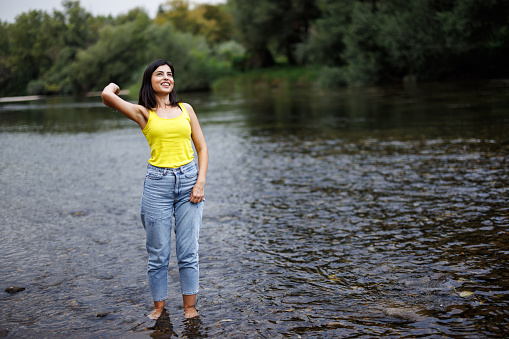 Young woman having fun while standing and skimming pebble into river on sunny summer day