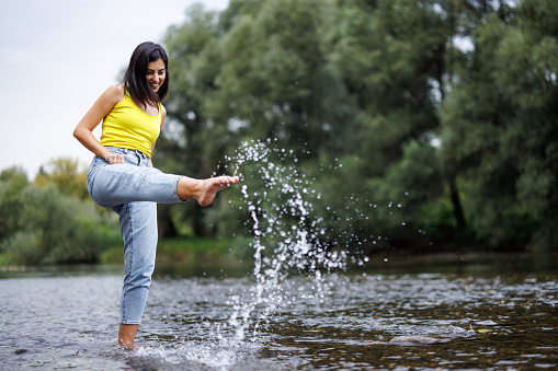 Young woman having fun while crossing river and splashing water on sunny summer day