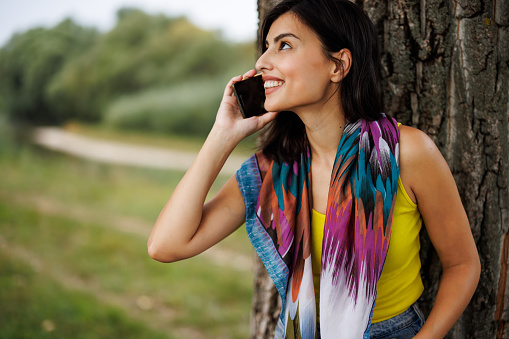 Beautiful young woman leaning on tree and talking on mobile phone in nature