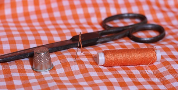 A spool of thread, thimble, needle and vintage scissors on orange and white checkered fabric