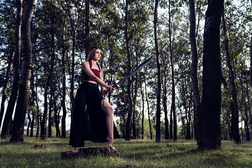 A young Caucasian girl with a top and a long skirt with deep cuts holding a sword in a forest