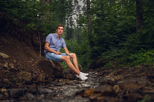 An attractive Caucasian white man sitting on a rock in the green forest at a small river