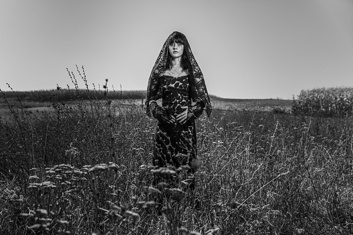 A grayscale of a young mourning woman with bangs in a black dress in a field