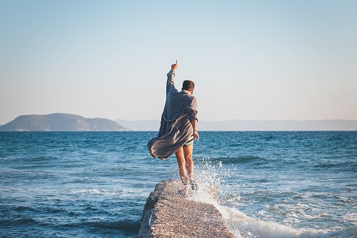 A Caucasian male in a bathrobe balancing in a cool pose on a rock splashed by the tide