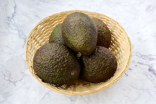 A top view of a wooden basket with avocados on a marble table