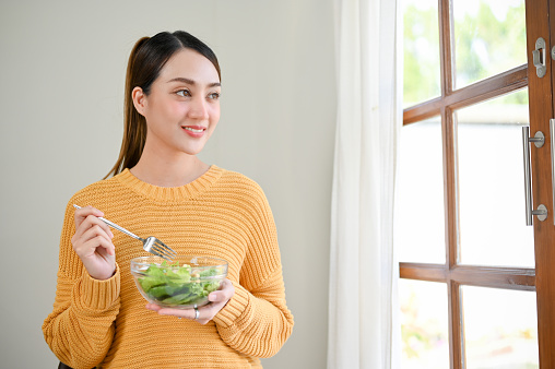 Attractive beautiful millennial Asian woman standing in the living room, holding a salad bowl, smiling, looking out the window, daydreaming about healthy life.