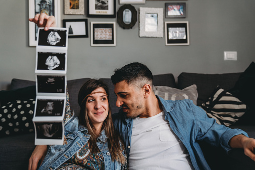 Millennial pregnant couple is holding a baby ultrasound together. They are lying down near to the sofa at home.