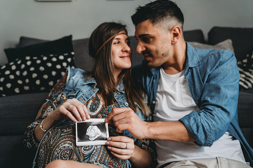 Millennial pregnant couple is holding a baby ultrasound together. They are lying down near to the sofa at home.