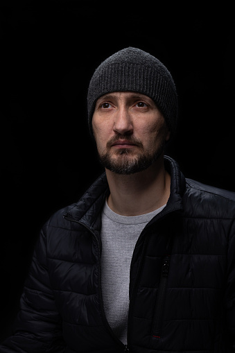 Portrait of a man with a beard on his face. The man is dressed in casual clothes.  The man wears a warm jacket and a hat on his head. The portrait of a man is photographed against a black background.