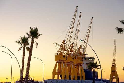 Huelva, Spain – July 15, 2020: Port of Huelva at sunset. Large yellow cranes and palm trees as the sun sets