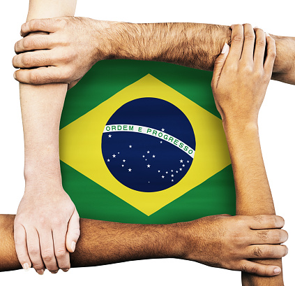 Multiracial group of hands and arms of men and women clasping each other, signifying unity, solidarity, cooperation and teamwork, around the Brazilian flag.