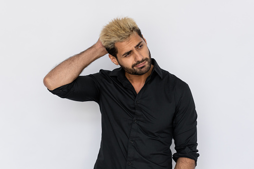 Portrait of doubtful young man scratching head over white background. Caucasian guy with stubble and highlighted hair wearing black T-shirt thinking or planning. Uncertainty and doubt concept