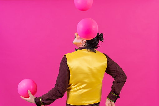 Clown with white facial makeup on a pink background, smiling juggling balls