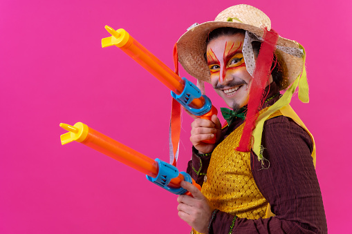 Clown with white facial makeup on a pink background, smiling with toy guns and hat