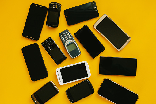 Flat lay depicting multiple generations of cell phone handsets on a yellow background.