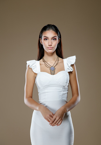 Portrait beautiful young fashion model with hair back wearing white dress with ruffles on shoulders, hand in hand in front and looking at camera, studio shot, beauty and fashion industry