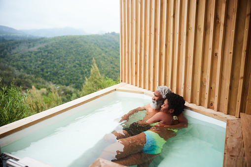 Mature couple wearing swimsuits laying together in a hot tub on a balcony and looking at the scenic view during a vacation