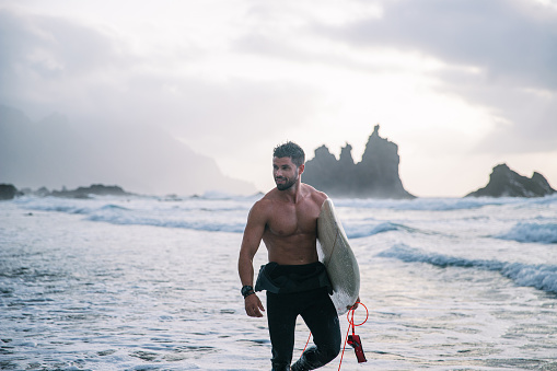 Surfer with surfboard walks on the beach, attractive guy looks around