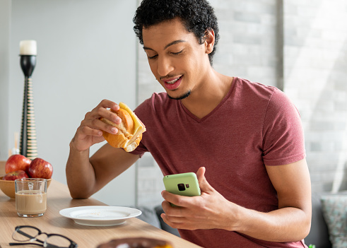 Brazilian man eats breakfast and looks at cell phone on sofa in living room at home