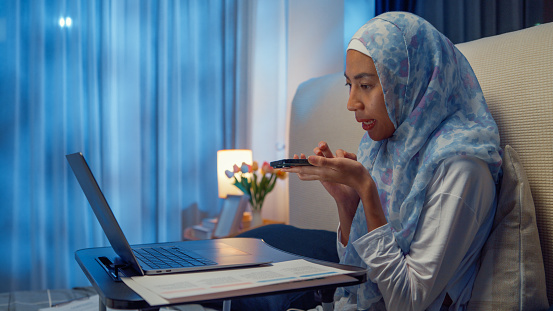 Happy young muslim woman sit on bed with computer laptop on folding laptop table and use smartphone giving voice commands to virtual assistant in bedroom at home at night. Work from home concept.