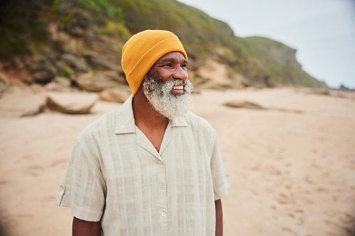 Smiling mature man with a gray beard wearing a knit hat while standing alone on a sandy beach on an overcast day