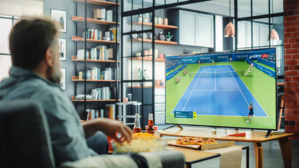 At Home Tennis Fans Sitting on a Couch Watch Game on TV, Cheer when Favourtite Sportswoman Team Scores a Ball. Screen Shows Professional Players During World Cup. Over the Shoulder en casa, los fanáticos del tenis sentados en un sofá ven el juego en la televisión, animan cuando el equipo favorito de las deportistas anota una pelota. la pantalla muestra jugadores profesionales durante la copa del mundo. sobre el hombro