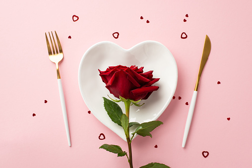 Valentine's Day concept. Top view photo of heart shaped dish with red rose knife fork and confetti on isolated pastel pink background