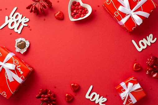 Valentine's Day concept. Top view photo of gift boxes with ribbon bows heart shaped dish with sprinkles chocolate candies inscriptions love you on isolated red background with copyspace in the middle