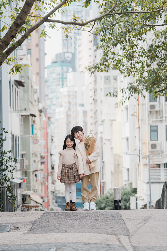 Full body shot of young smiling fashionable children, Asian boy and Asian girl, who are teenage friends wearing autumn or winter clothes, holding hands and bunch of flowers, standing next to each other on the street in the city with skyscrapers and trees. They are looking at camera.

Young Asian brother and sister, spending time together on the streets as family siblings during weekend or free time, enjoying the day in Autumn.