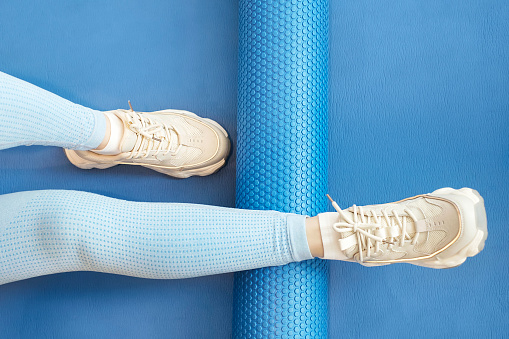 Female leg rolls a massage foam roller close-up on a blue background, top view.