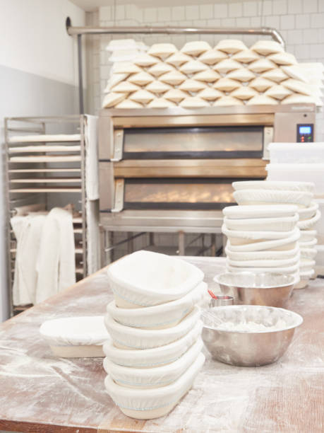 istock scene inside a bakery: work table for kneading bread with stacked bread pans and flour bowls, ovens and cooling rack trolley behind 1459663131