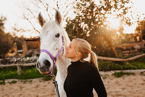 Portrait of the friendship between a blond young woman and her white mare in a rustic stable outdoors in Majorca. Real life. Color editing. Part of a series.