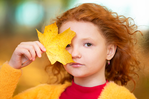 Red-haired little girl closes one eye with a yellow maple leaf. Sad child on an autumn day close-up