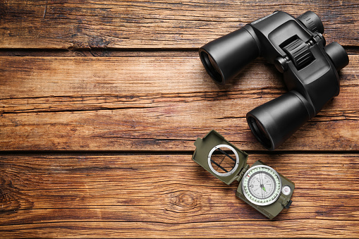 Binoculars and compass on wooden table, flat lay. Space for text
