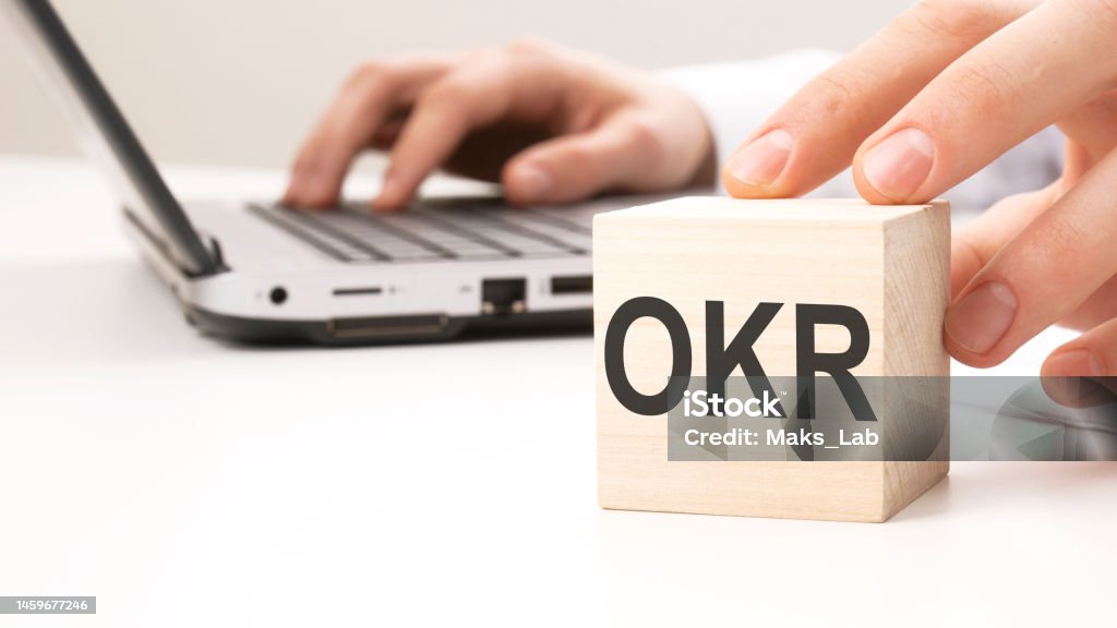 OKR text wooden block on white table background. Idea, strategy, advertising, marketing, keyword and content concept man holding wooden cube witt letters OKR in the background a laptop on a white office table background Finance Stock Photo