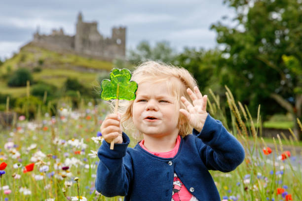 배경에 Cashel 성의 바위가 있는 아일랜드 클로버리프 롤리팝을 가진 귀여운 유아 소녀. 건강에 해로운 과자를 먹는 꽃 초원에서 행복한 건강한 아이. 아일랜드의 가족 및 어린 자녀 휴가 스톡 사진