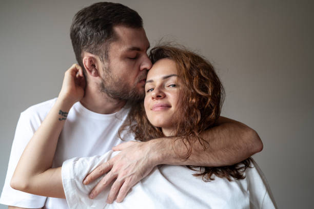 retrato de una pareja joven feliz vestida con ropa blanca abrazando y besando a una mujer de cabello rizado sobre un fondo liso. - heterosexual couple fotografías e imágenes de stock