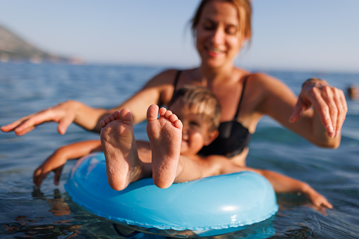 Happy young mother with blond hair plays with her little cheerful toddler son and rides him on a bright blue inflatable ring in the deep blue shiny Adriatic Sea