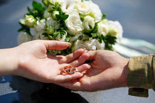 The hand of a guy in military uniform holds the hand of a girl with wedding rings against the background of a wedding bouquet of white roses