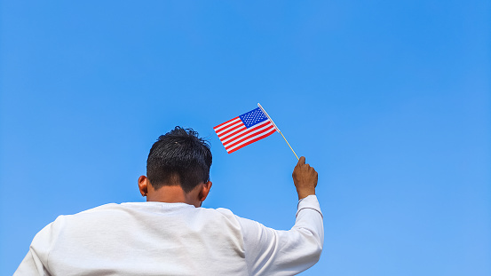 Boy holding US, USA or United States flag against clear blue sky. Man hand waving American flag view from back, copy space for text