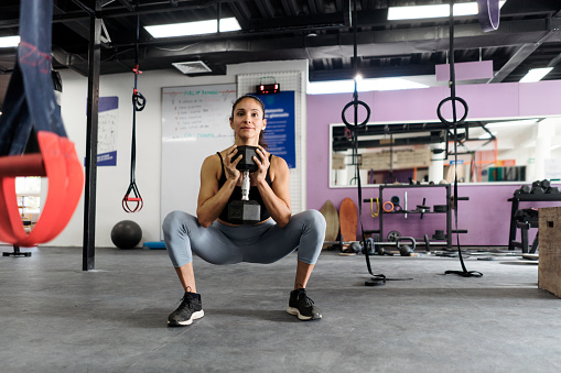 A young latin woman doing a squat at the gym and holding a dumbbell.