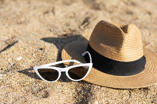 Close up hat and white trendy glasses on the sandy beach are not people. Straw fashionable hat with black ribbon. Concept: rest and vacation, travel agency. Sunny hot weather. Beige sand