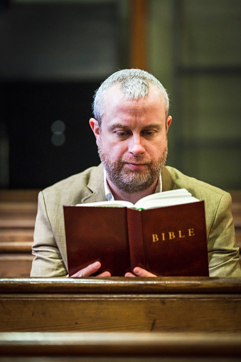 Close up of a mid adult Christian man reads the holy bible while sitting on a wooden pew in a church.