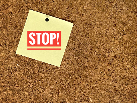 Close-up of red colored Stop! text on yellow sticky notepad pinned on cork board